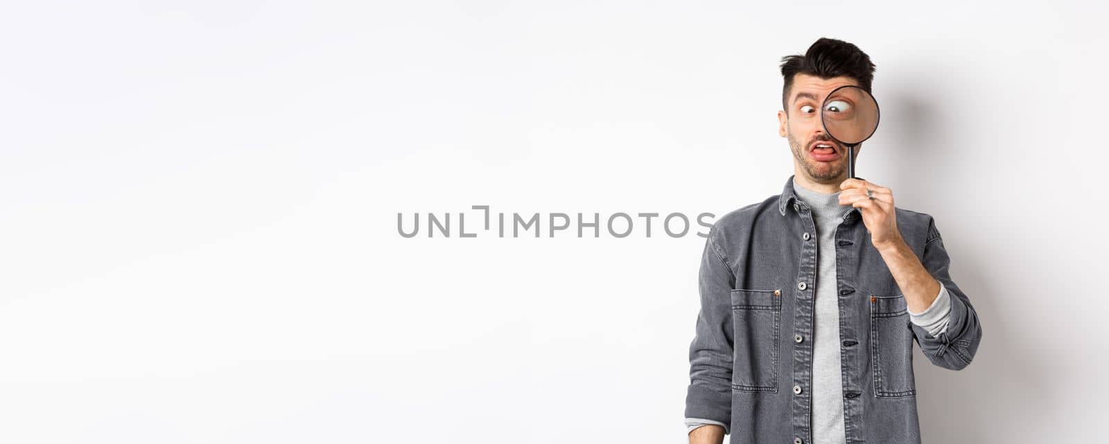 Funny young man squinting, showing faces with magnifying glass, standing on white background.