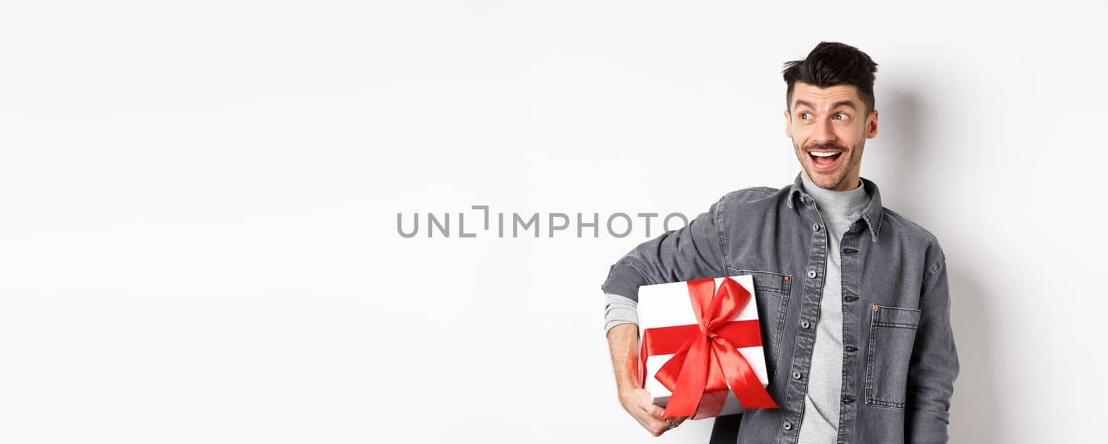 Cheerful handsome guy looking at empty space with amazed smile, checking out special valentines day offer, holding big romantic gift box, standing on white background by Benzoix