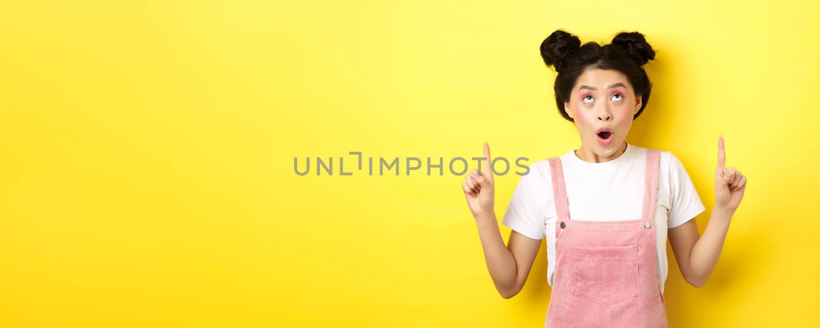 Excited asian teen student with glamour makeup, gasping amazed pointing fingers up, showing advertisement, yellow background.