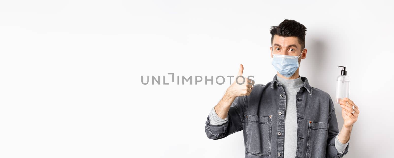 Healthy people and covid-19 concept. Excited man in sterile medical mask holding bottle of good hand sanitizer, show thumb up, recommend antiseptic, standing against white background.