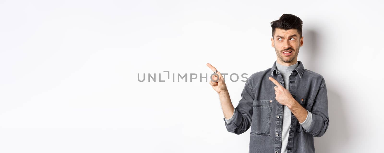 Skeptical and doubtful young man pointing fingers left at logo, look with dislike and confusion, standing on white background.