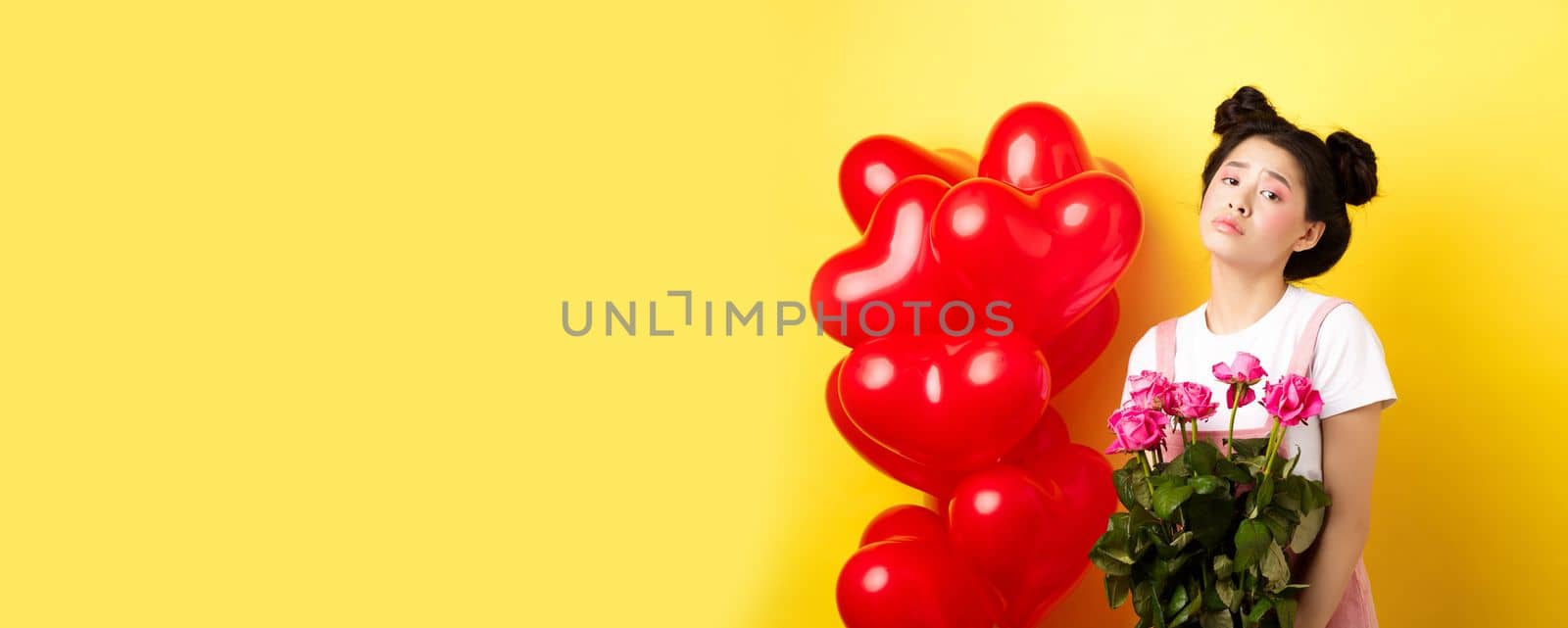 Happy Valentines concept. Sad and gloomy asian woman holding bouquet of roses and feeling upset and lonely on romantic lovers day, standing near red heart balloons, yellow background.