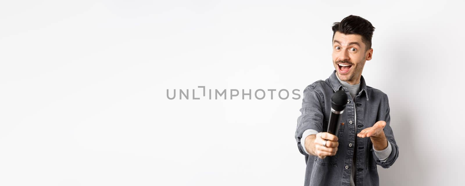 Handsome smiling man stretch out hand with microphone, giving you mic, standing on white background.
