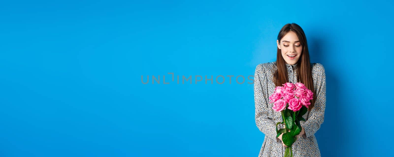 Valentines day concept. Happy attractive woman receive surprise flowers, looking thankful at bouquet of pink roses, standing on blue background by Benzoix