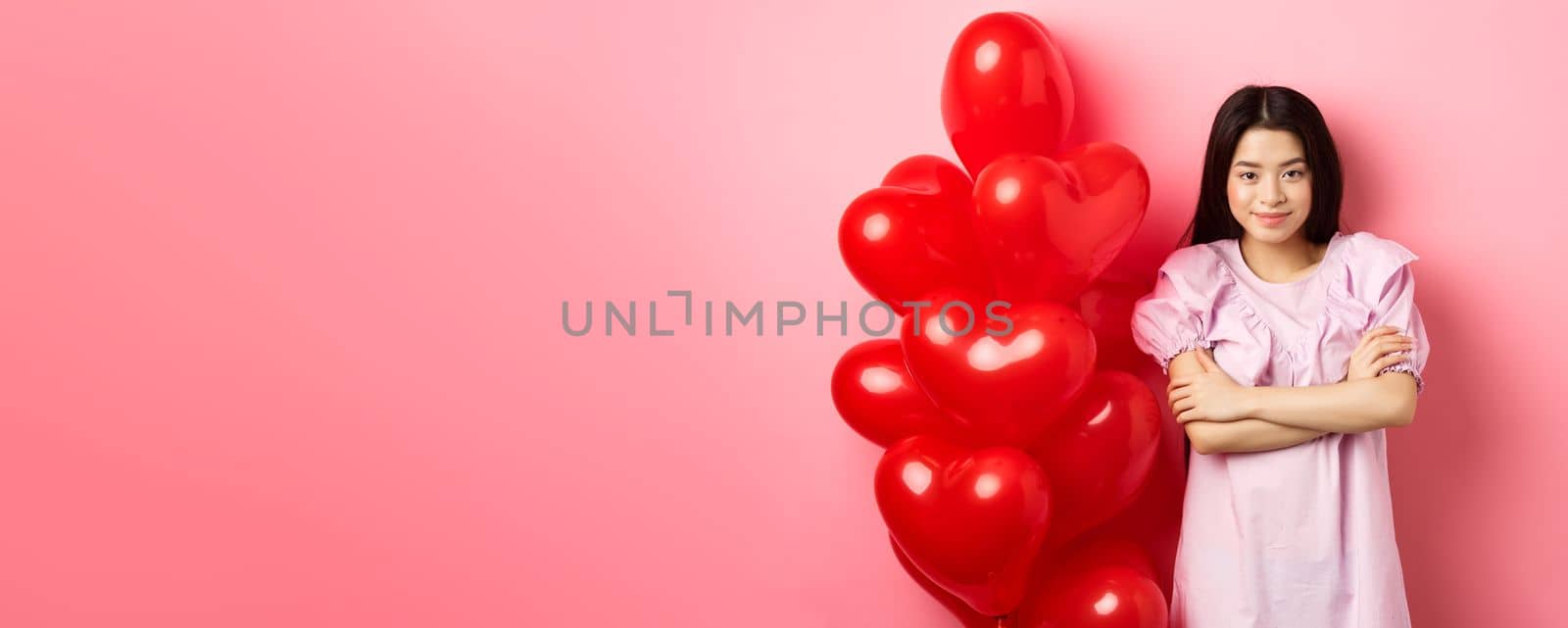 Confident teenage girl cross arms on chest and smile, celebrating valentines day in cute dress with red heart balloons, standing on pink background by Benzoix