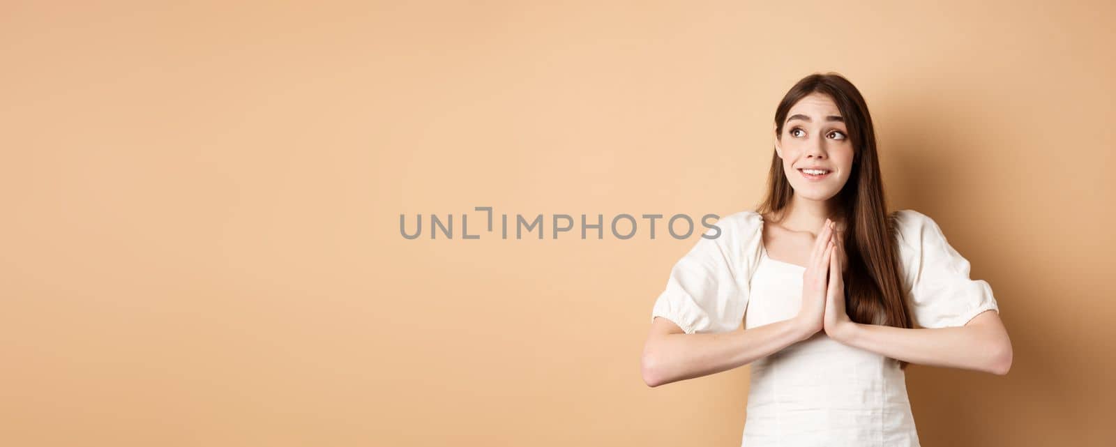 Hopeful young girl begging god and looking dreamy at upper left corner, praying or making wish, standing on beige background.