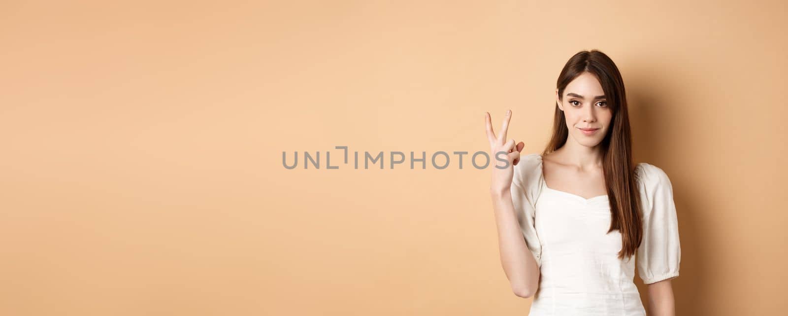 Attractive young woman show fingers number two, smiling and looking confident, standing on beige background by Benzoix
