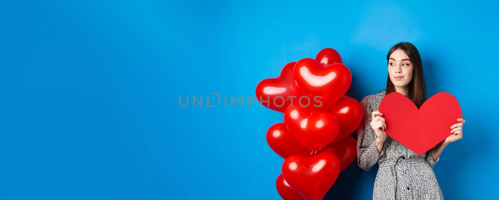 Valentines day. Romantic girl in dress standing near balloons and holding big red heart cutout, dream of something, standing on blue background.