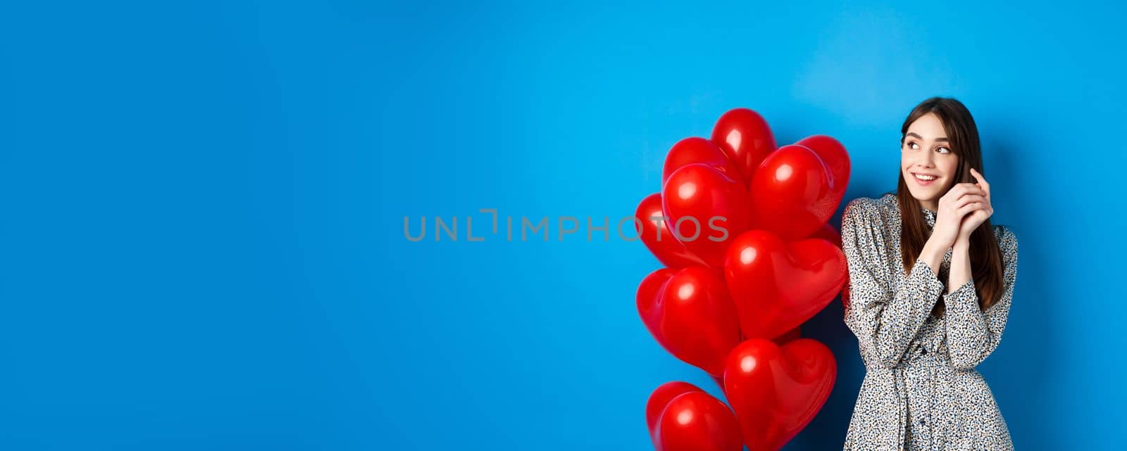 Valentines day. Romantic young woman in dress, looking dreamy left and smiling, standing near red hearts balloons, blue background by Benzoix