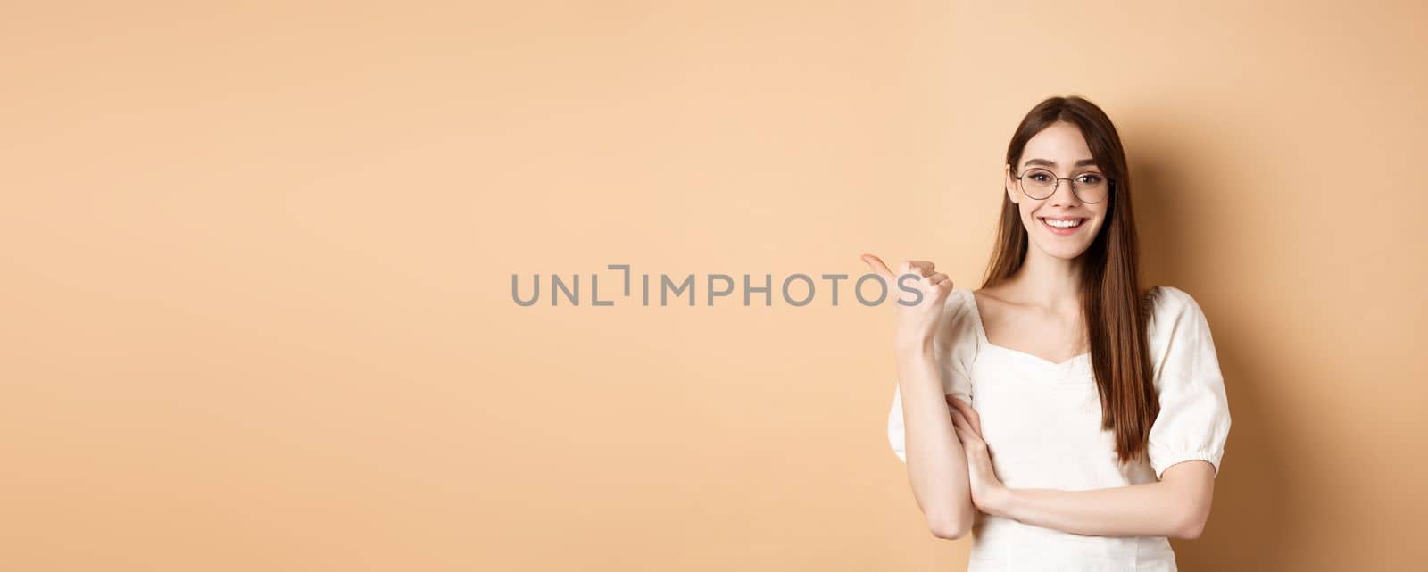 Cheerful woman in eyewear pointing finger left at logo, smiling happy, standing on beige background by Benzoix