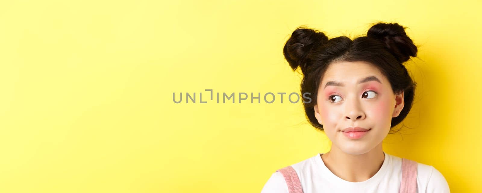 Close-up portrait of cute glamour asian girl, bright makeup and hairstyle, looking aside at logo with silly face, yellow background.