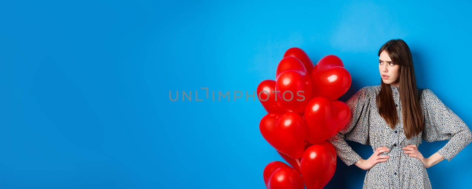 Valentines day. Annoyed and bothered girl complaining, looking left at empty space and frowning, standing near heart balloons, blue background by Benzoix
