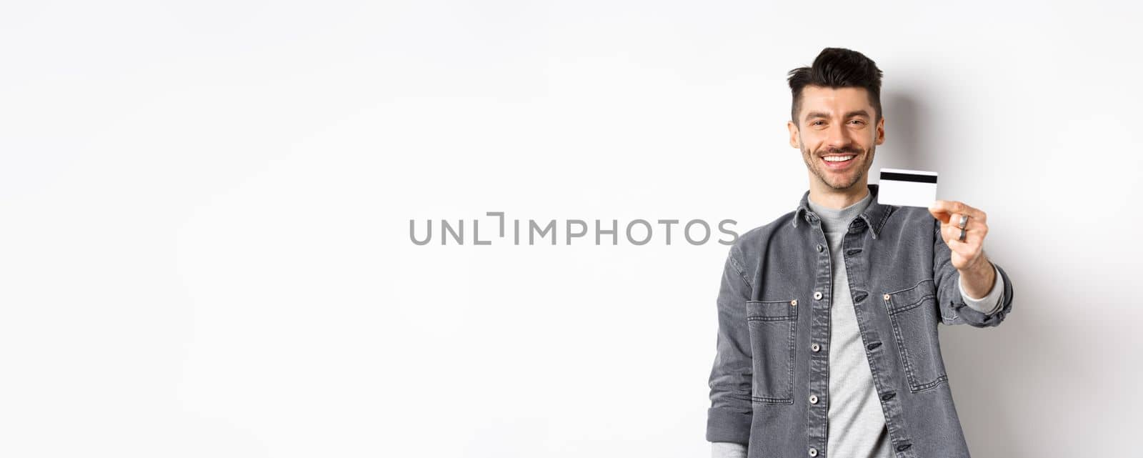 Handsome stylish guy showing plastic credit card and smiling, satisfied client standing on white background.