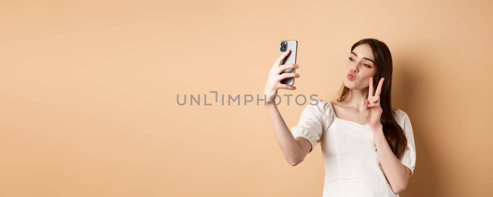 Fashion female blogger pucker lips and show v-sign at smartphone camera, taking selfie for social media, standing on beige background.