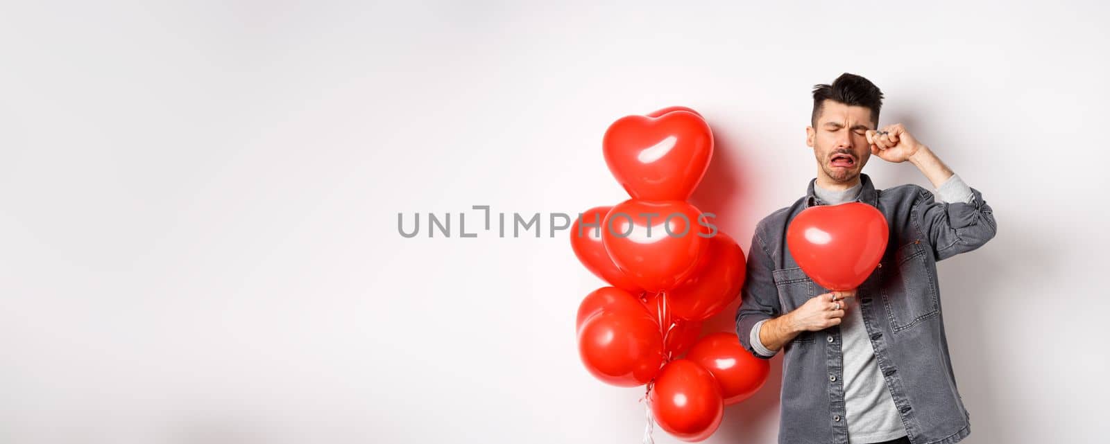 Valentines day and love concept. Sad crying man holding red heart balloon and whiping tears, standing single and miserable, being heartbroken, white background.