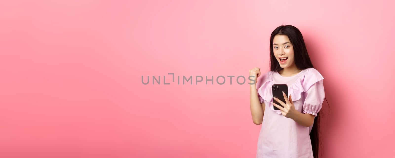 Online shopping. Satisfied asian girl winning on mobile phone, say yes and make fist pump, holding smartphone, standing against pink background by Benzoix