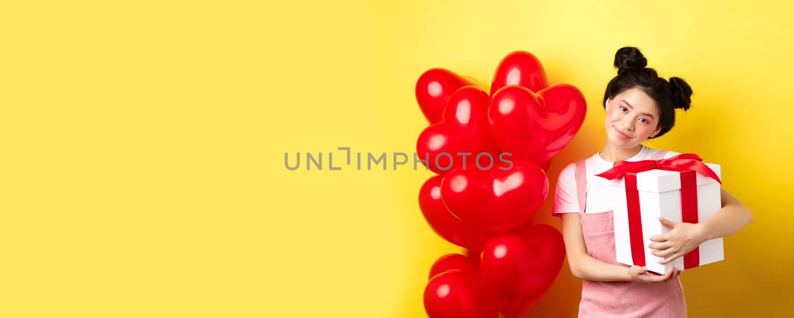 Happy Valentines day. Cute asian girl hugging surprise gift from boyfriend, standing near lovely red heart balloons and yellow background.