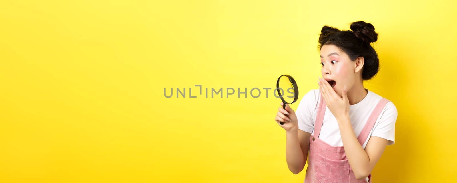 Excited silly asian girl gasping wondered, found something interesting, looking through magnifying glass, standing on yellow background by Benzoix