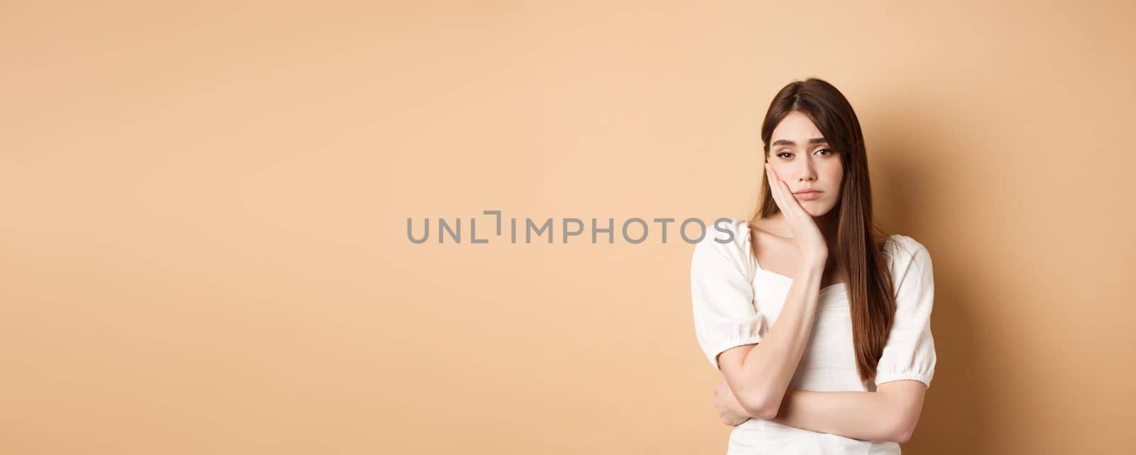 Bored and tired young woman looking indifferent at camera, lean face on palm and stare at camera unamused, standing sleepy on beige background.