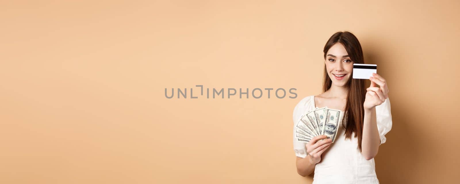 Smiling girl showing platic credit card to camera, holding dollar bills close to chest, standing on beige background by Benzoix