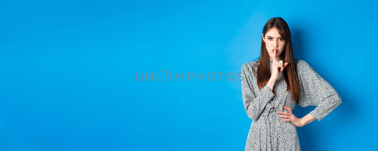 Angry and bossy young woman tell to keep quiet, show shush taboo sign and frowning upset with bad behaviour, standing on blue background by Benzoix