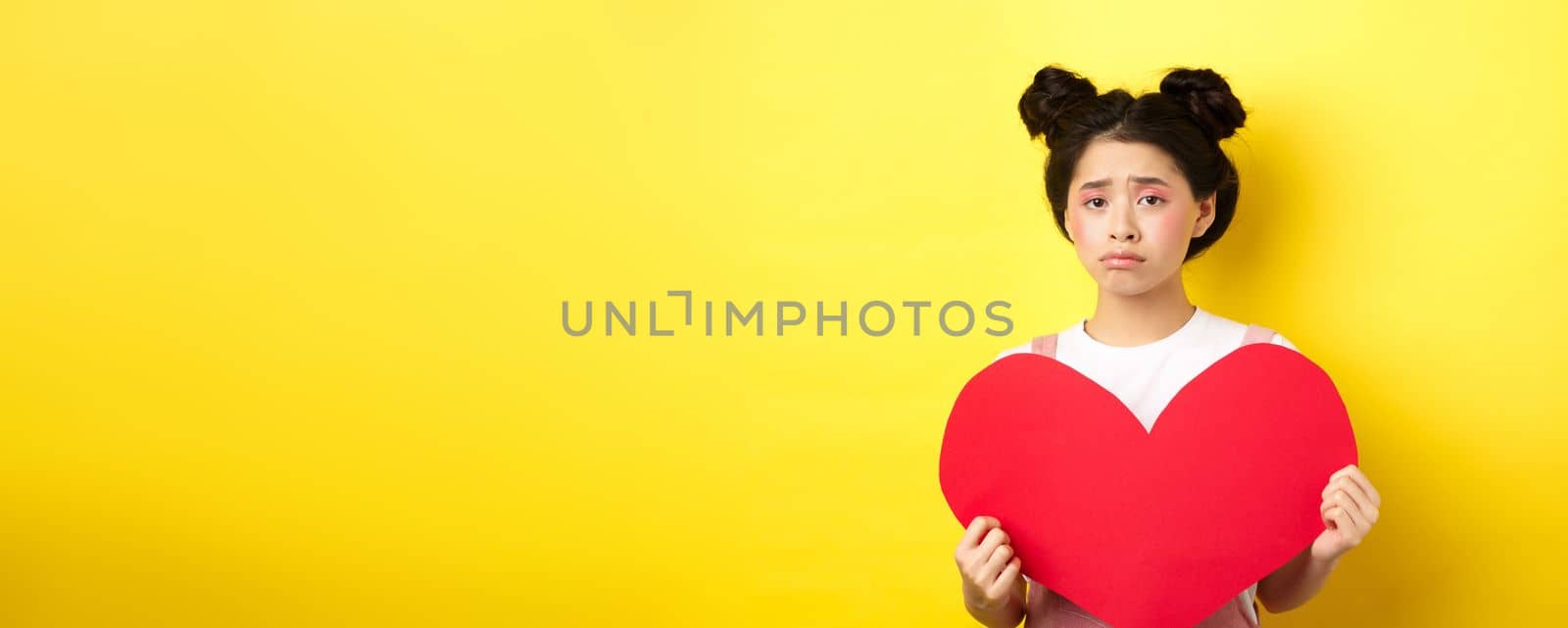 Heartbroken asian woman showing big red heart cutout and looking sad, feeling lonely on lovers day, showing valentine card at camera, yellow background.
