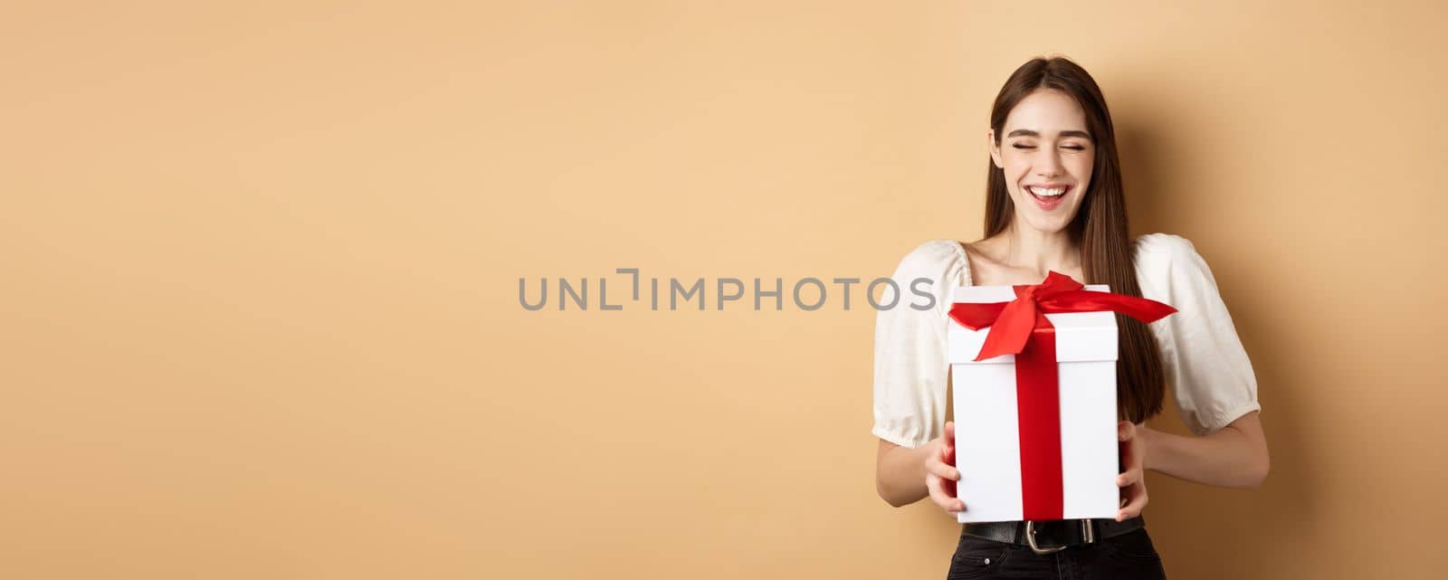 Happy smiling woman laughing, holding gift box from lover, enjoying romantic date on Valentines day, standing on beige background by Benzoix