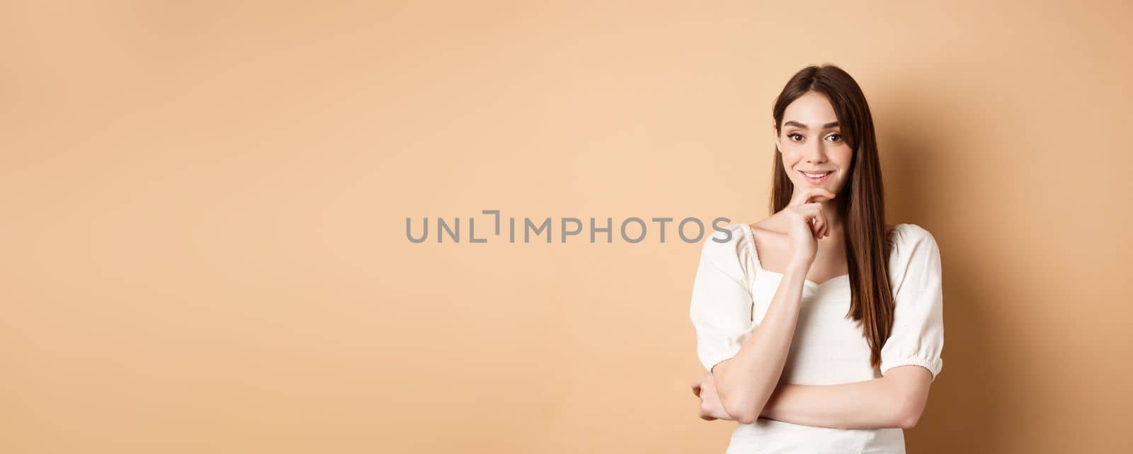 Smiling young european woman in dress, having an idea, listening with interest, touching chin and looking at camera intrigued, standing against beige background.