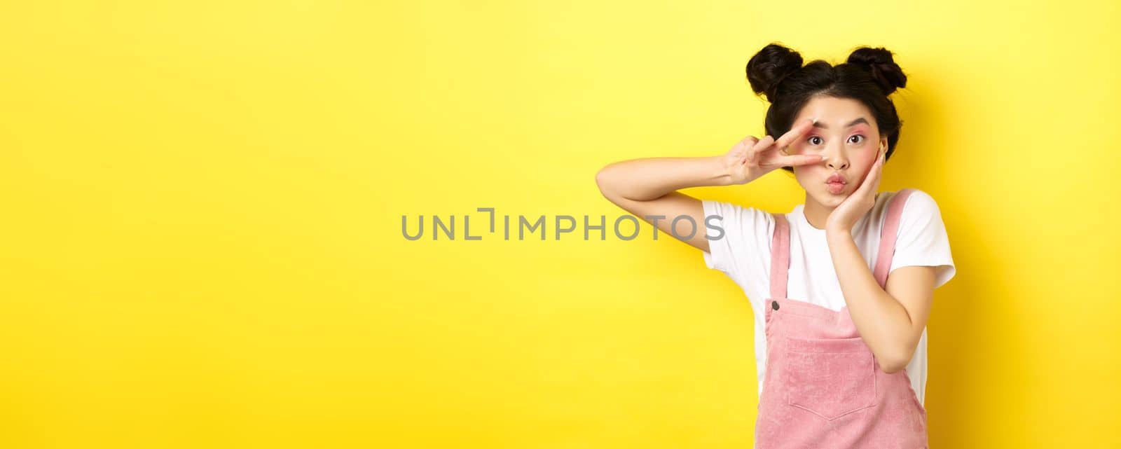 Kawaii asian girl showing v-sign and pouting cute, making silly face with makeup, standing on yellow background by Benzoix