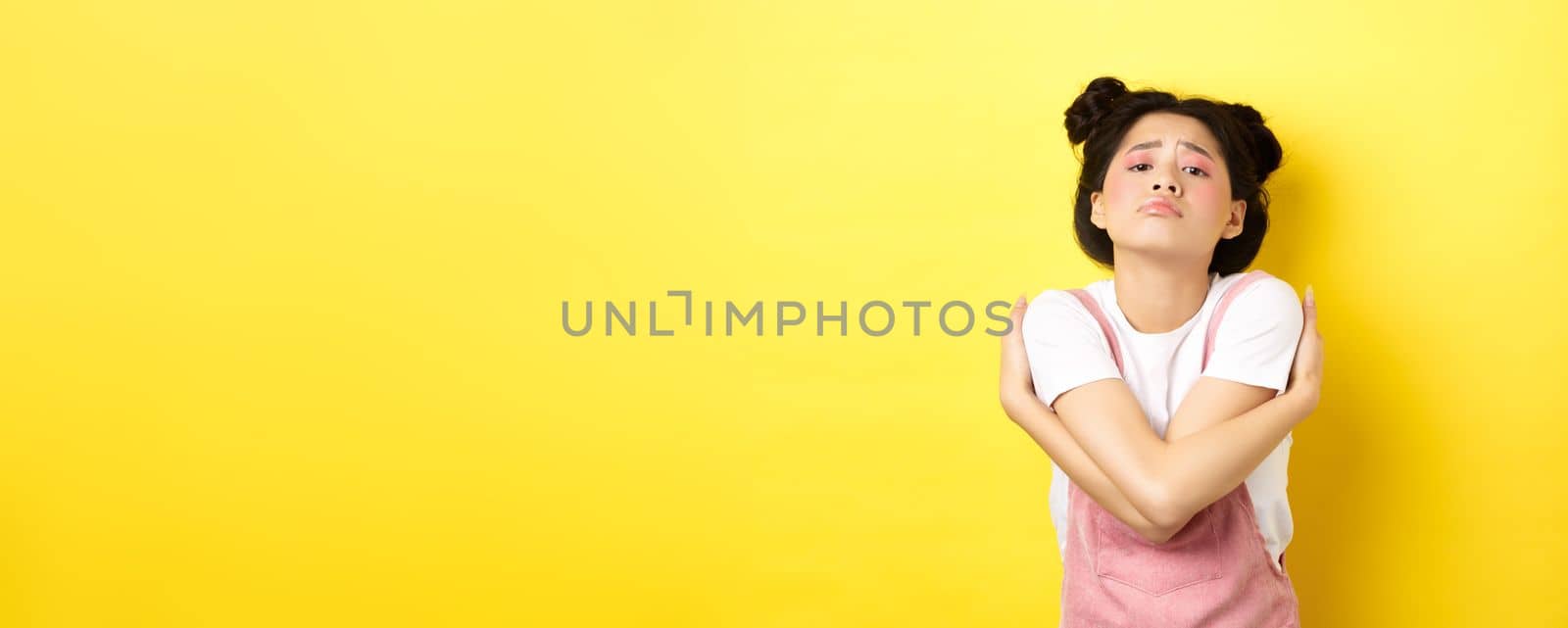 Lonely sad teen girl hugging herself, wanting to relationship and cuddles, standing alone on yellow background.