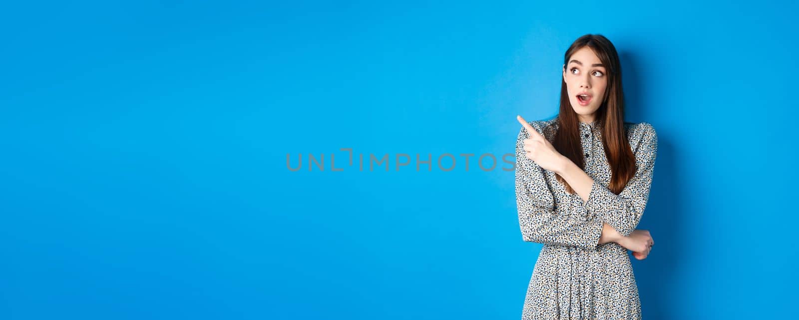 Wow look there. Excited pretty lady in dress looking and pointing left wondered, checking out advertisement, standing on blue background by Benzoix