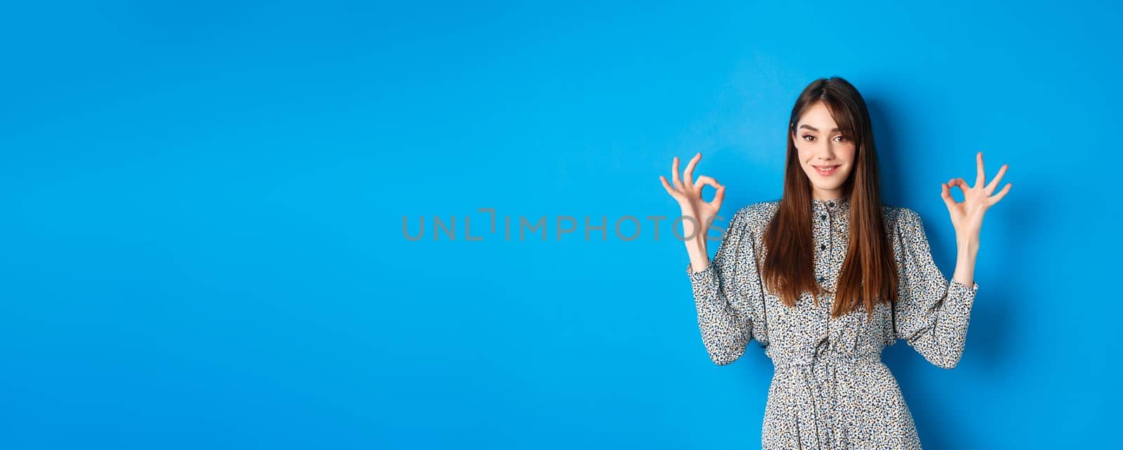 Confident adult woman in dress with natural hair color, showing okay signs and smiling, like and approve something good, standing on blue background by Benzoix