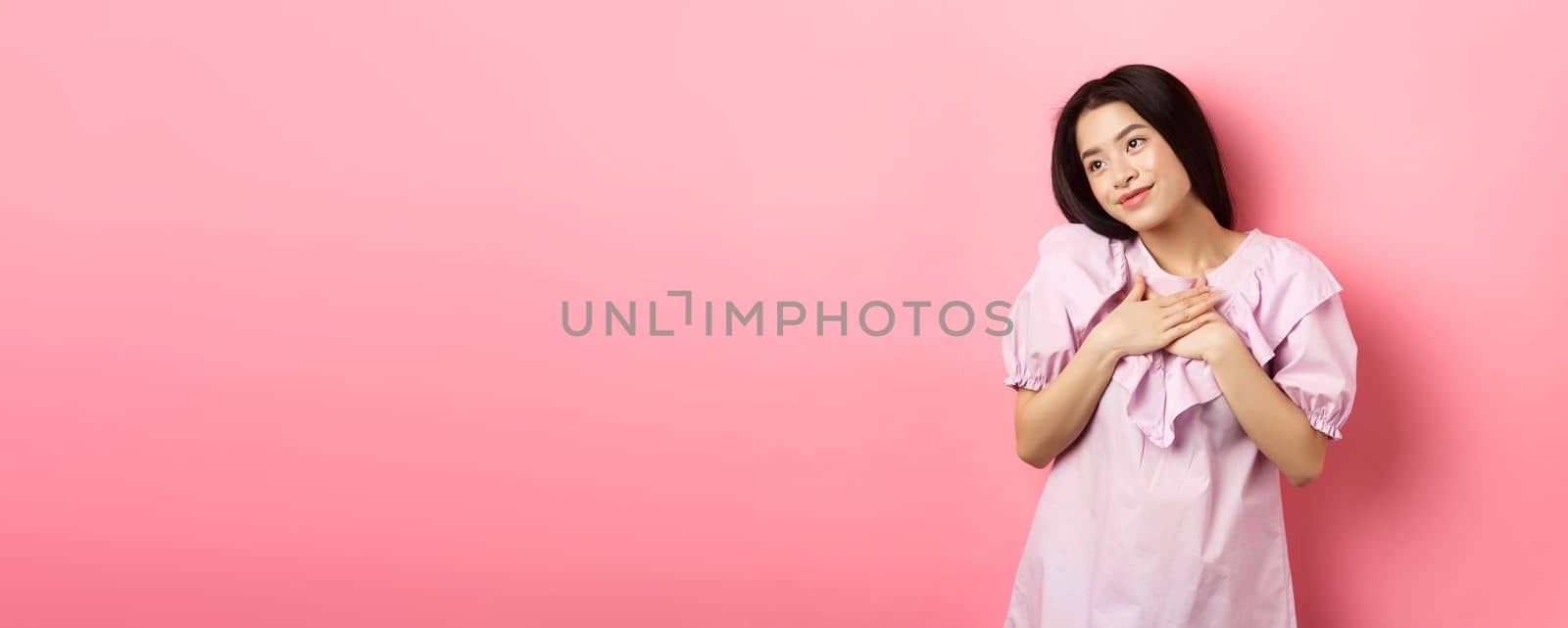 Dreamy asian girl smiling and holding hands on chest, looking left at something romantic and cute, standing on pink background by Benzoix