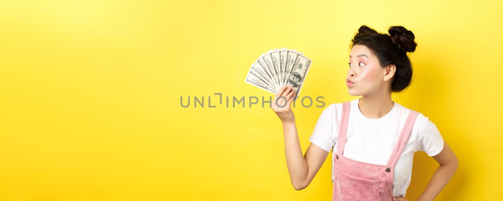 Shopping. Silly korean girl with glam makeup, pucker lips and looking at dollar bills, wasting money, standing on yellow background by Benzoix