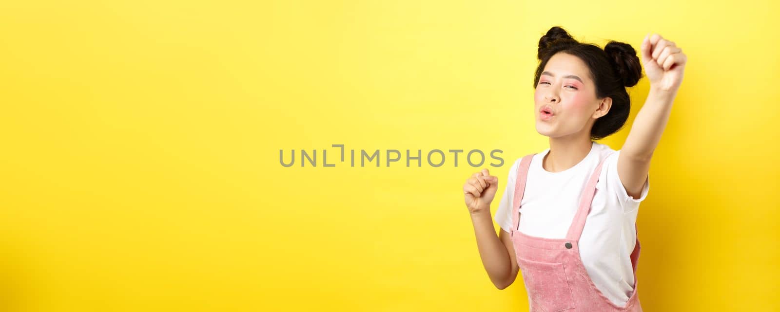 Excited asian girl looking motivated, raising hand up and chanting, cheering with joy, standing happy on yellow background.