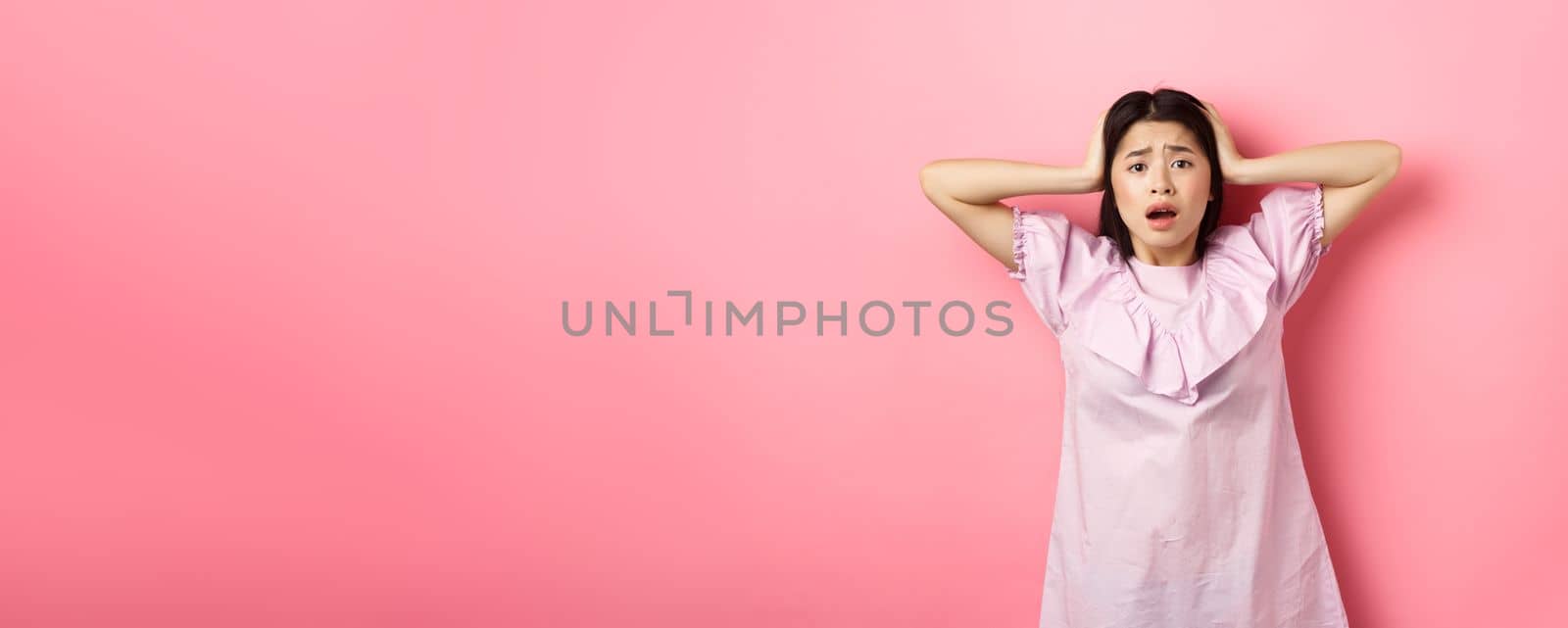 Frustrated asian teen girl panicking, holding hands on head and screaming scared, standing anxious on pink background.