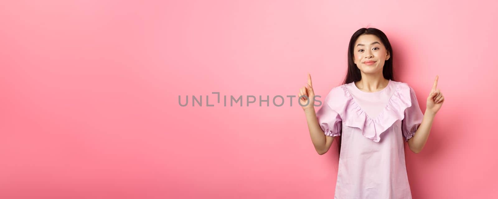 Beautiful young asian woman in romantic dress pointing fingers up, smiling and gladly showing advertisement, standing on pink background by Benzoix