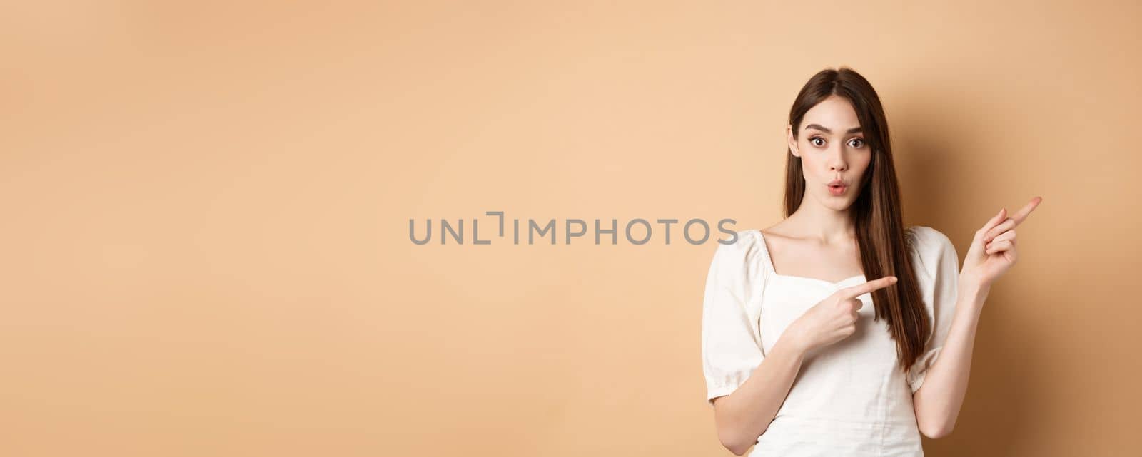 Wow look here. Impressed caucasian woman in white dress pointing fingers right and look curious, asking question about product, beige background by Benzoix