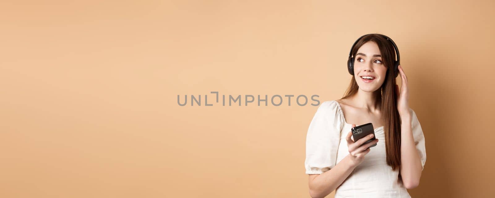 Attractive girl listening music in wireless headphones, holding mobile phone and looking aside with dreamy smile, beige background.