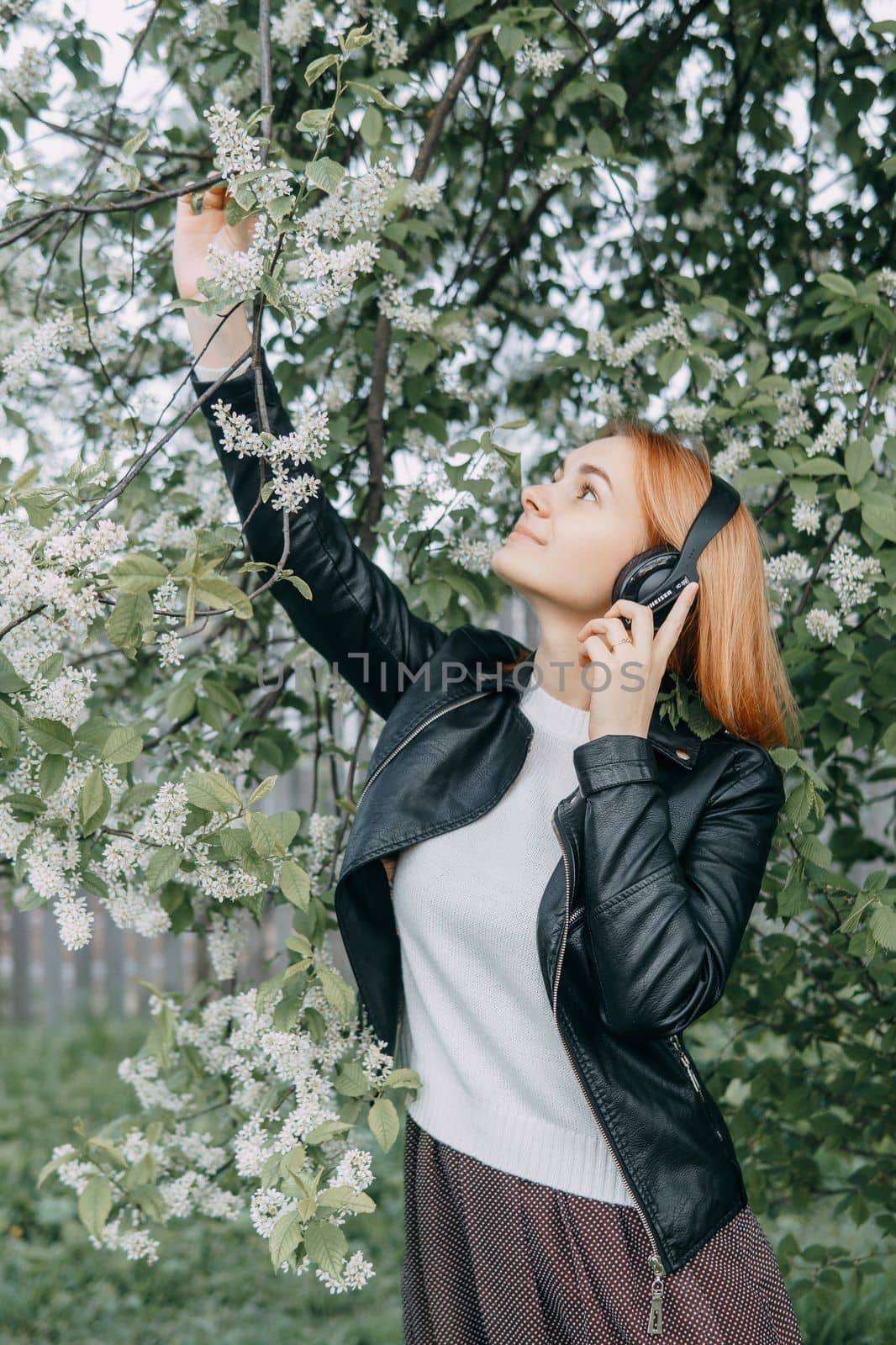 A red-haired teenage girl listens to music in headphones, configured via the phone. The time of spring flowering of cherry orchards.