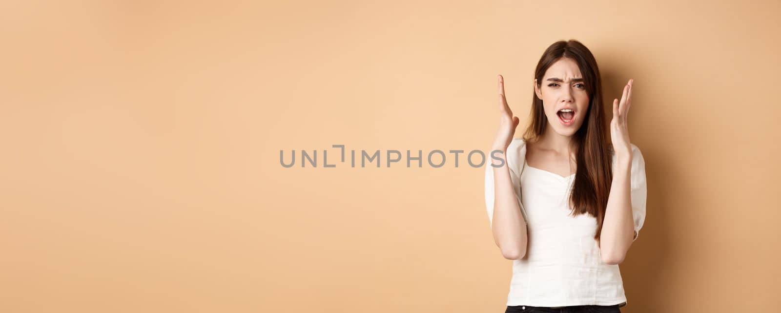 Frustarted woman shouting and shaking hands angry, looking mad, standing distressed on beige background by Benzoix