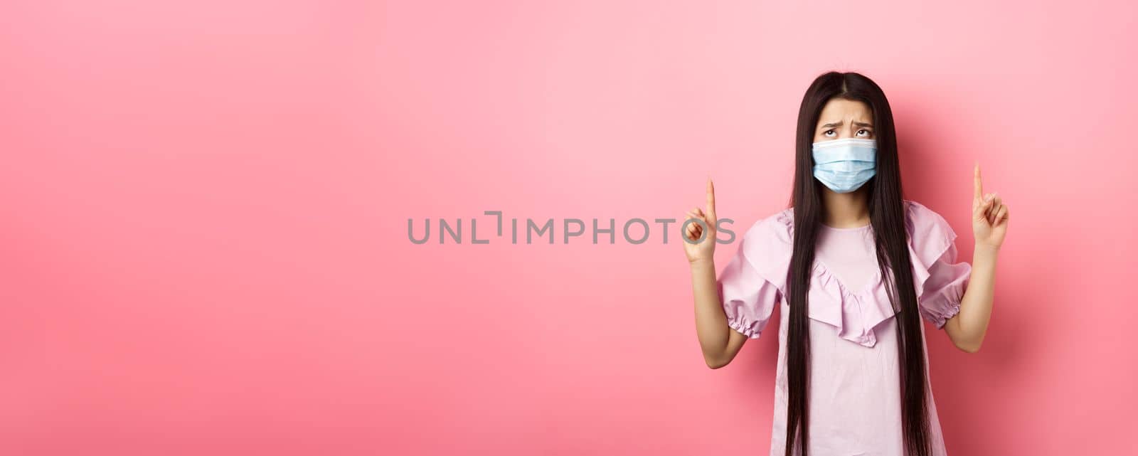 Coronavirus, quarantine and lifestyle concept. Disappointed sad asian girl pointing fingers up, frowning and sulking at unfair situation, standing against pink background by Benzoix