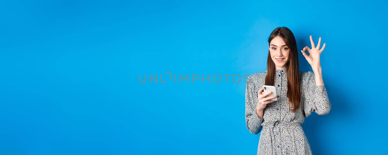 Beautiful young woman in dress showing okay sign and using smartphone, smiling at camera, standing on blue background.