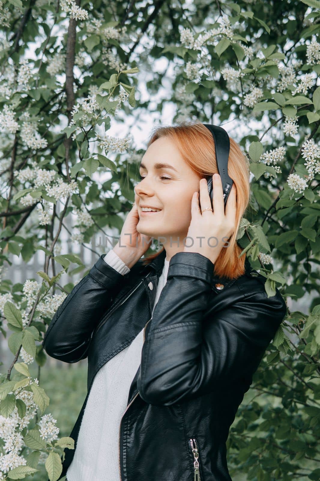 A red-haired teenage girl listens to music in headphones, configured via the phone. The time of spring flowering of cherry orchards.