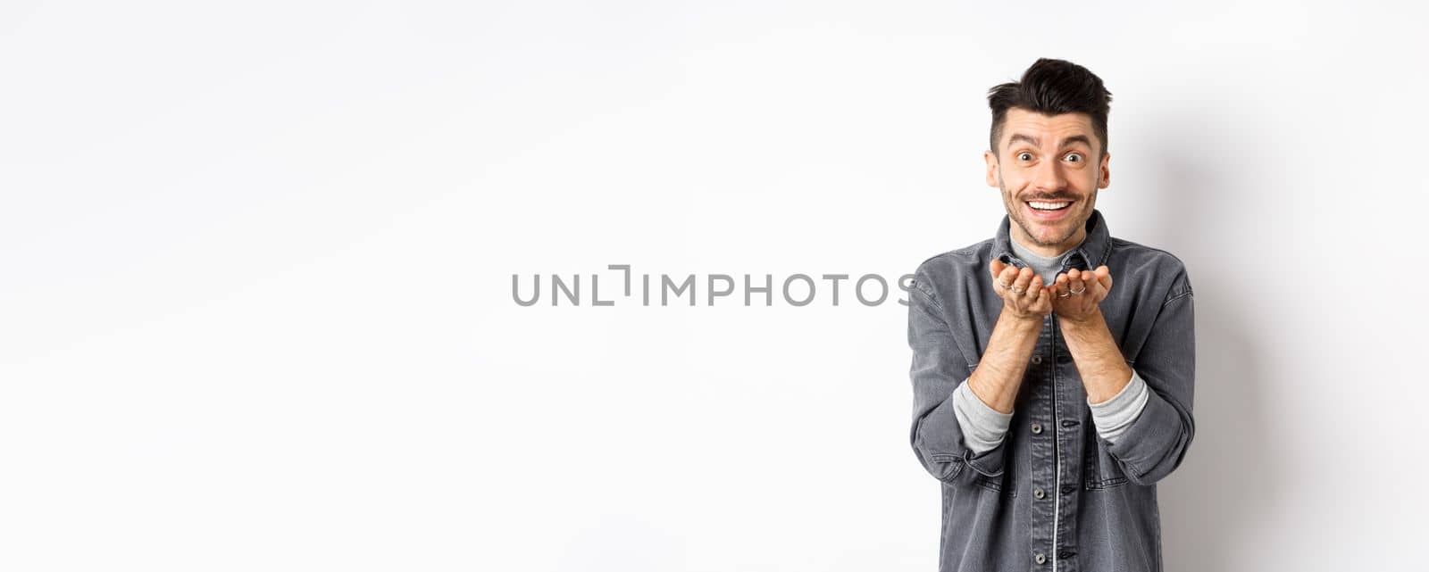 Lovely young man sending air kiss at camera and smiling. Hopeless romantic showing his love, holding something in hands, standing on white background.