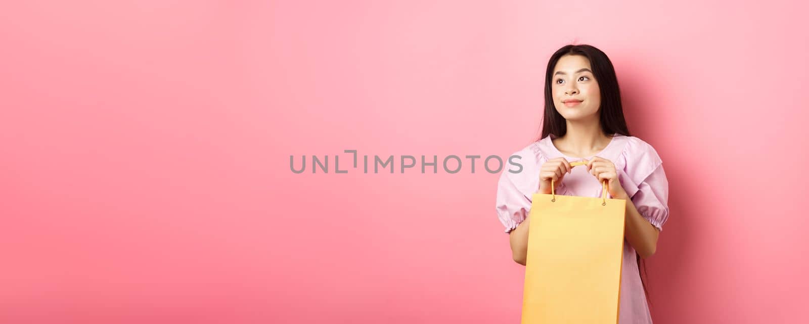 Beautiful asian girl dreaming and holding shopping bag, standing on pink background.