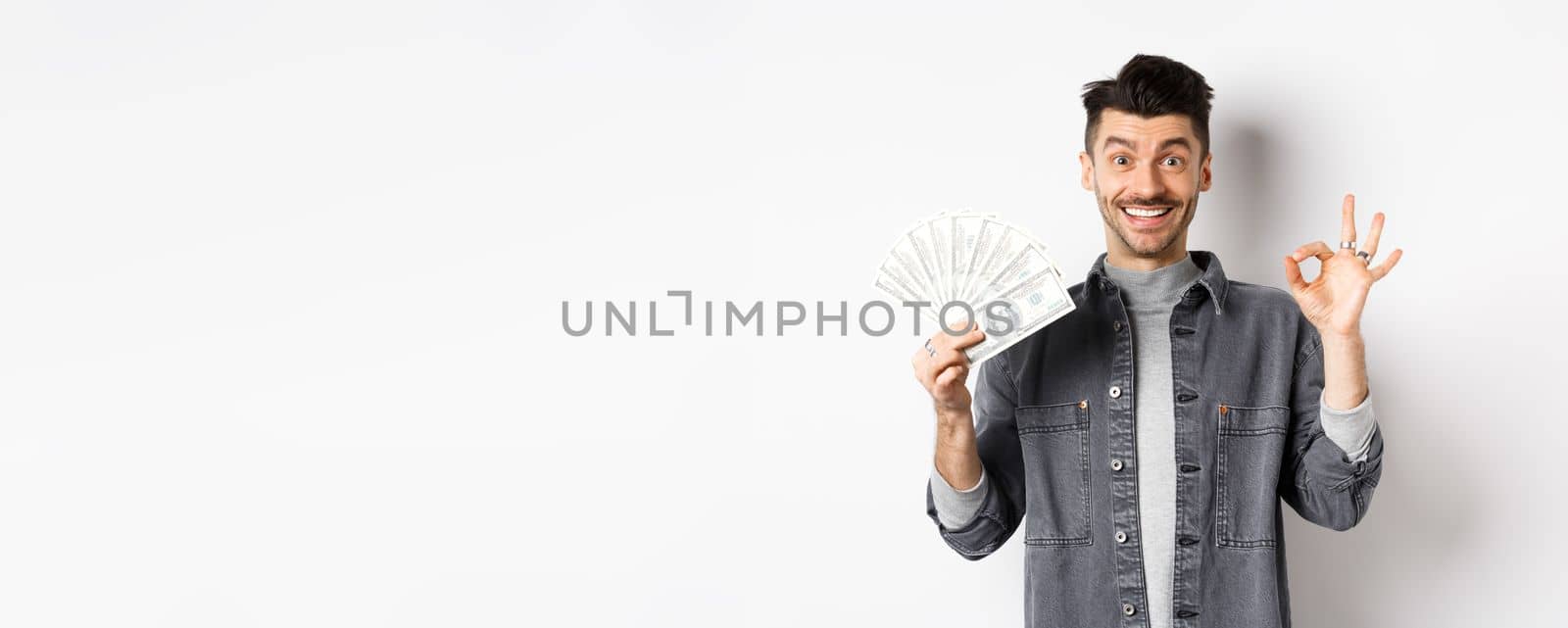 Image of excited man holding dollar bills and show okay sign with happy face, making money, standing on white background by Benzoix