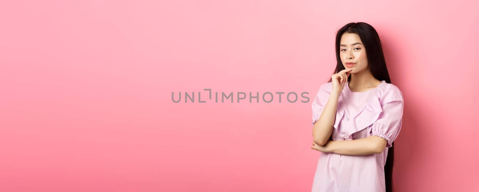 Hmm, interesting. Pensive and cunning asian girl having idea, plan to do something, looking at camera and touching chin with thoughtful face, standing on pink background.