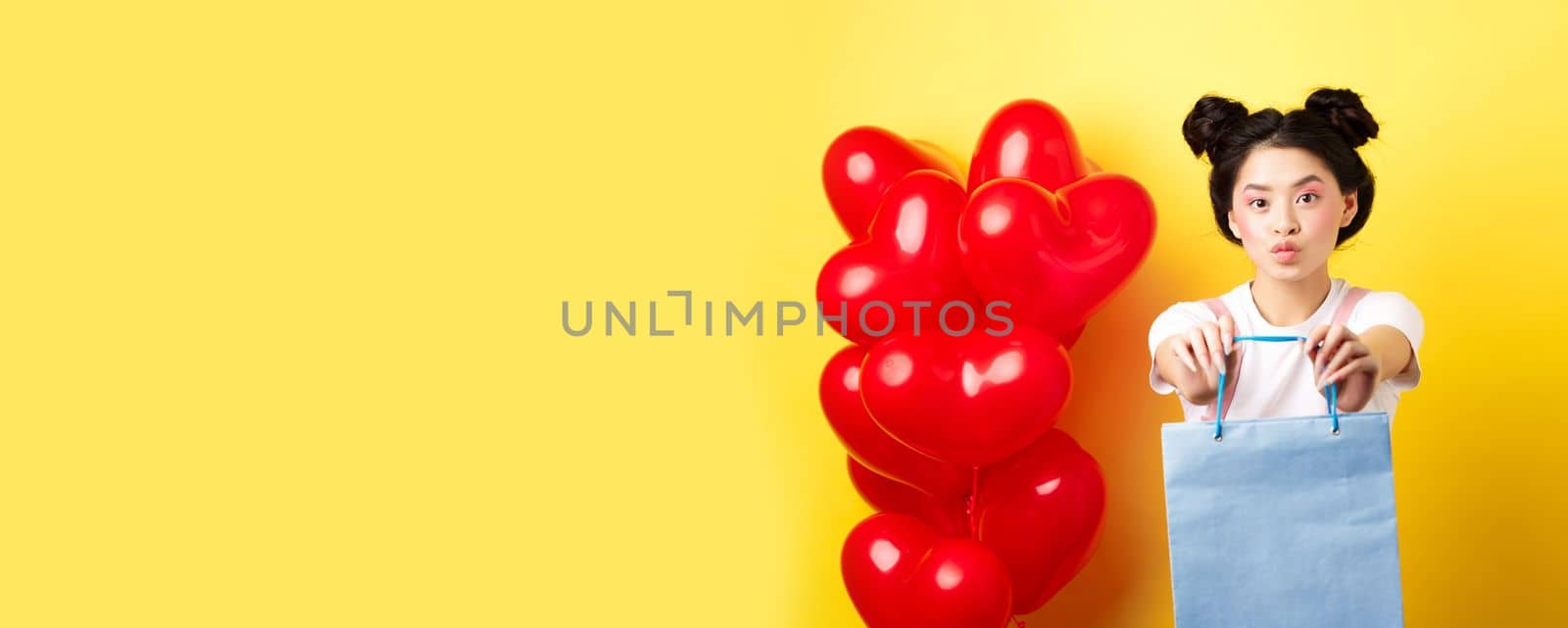 Happy Valentines day. Cute asian girlfriend giving lover a gift, stretch out hands with shopping bag and looking silly, standing over yellow background near heart balloons.