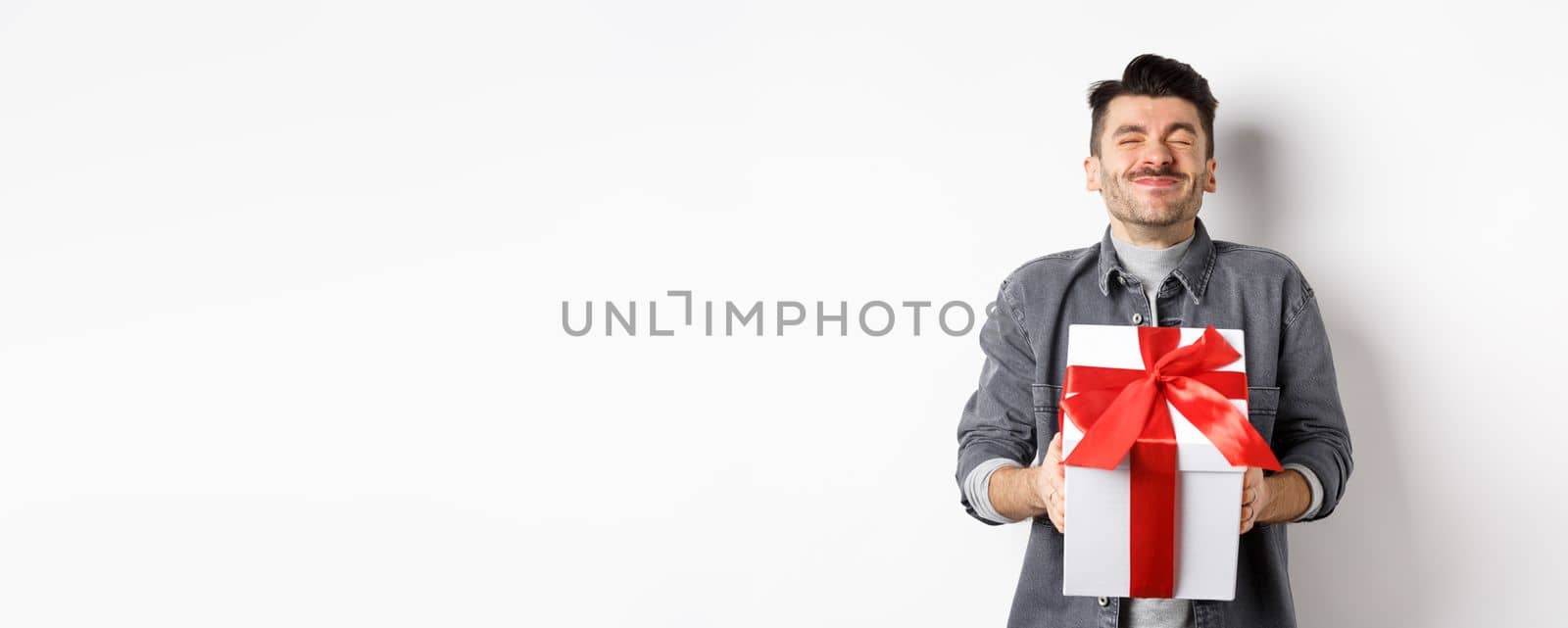 Happy handsome young man holding big valentines day gift and cheering, receive romantic surprise present from lover, standing on white background.